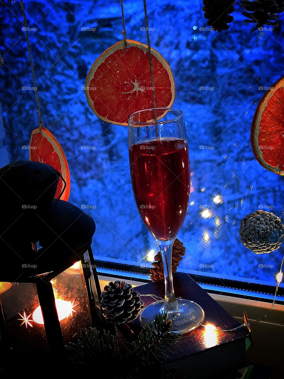 A glass of red wine stads on the windowsill. Next to it is a lantern with a luminous candle and pine cones. Dried oranges and cones hang on the window. Outside the window trees in the snow