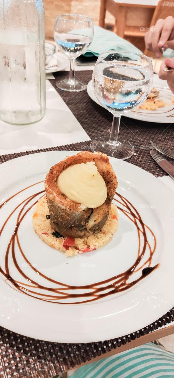 Baked fish with mashed potatoes and couscous in a plate on a table with drinks on the terrace of a street restaurant on a summer day in italy, close-up side view. Sea food concept.