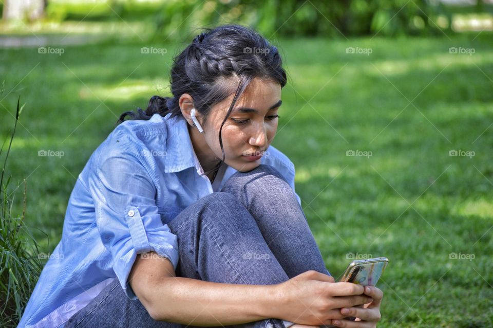 a girl in jeans and a shirt sits on a lawn in a park with a phone in her hands