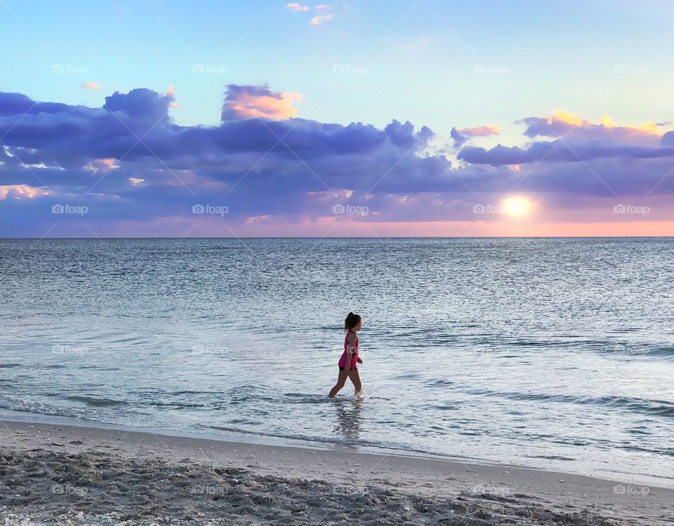 Purple clouds over a golden ocean sunset surrounding a little girl playing in the waves.