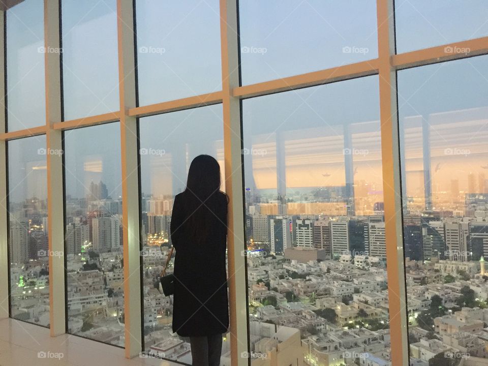 Woman dreaming looking out into the city of Abu Dhabi from a high floor in a skyscraper with panoramic views of the architecture. Conquer the world.