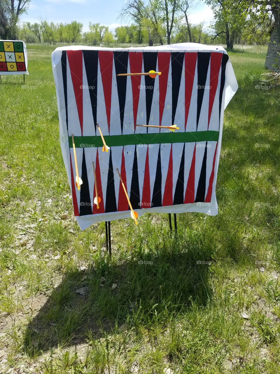 An archery target designed to look like a backgammon board with arrows peppering the board.   The target stands in a field on a beautiful sunny day.