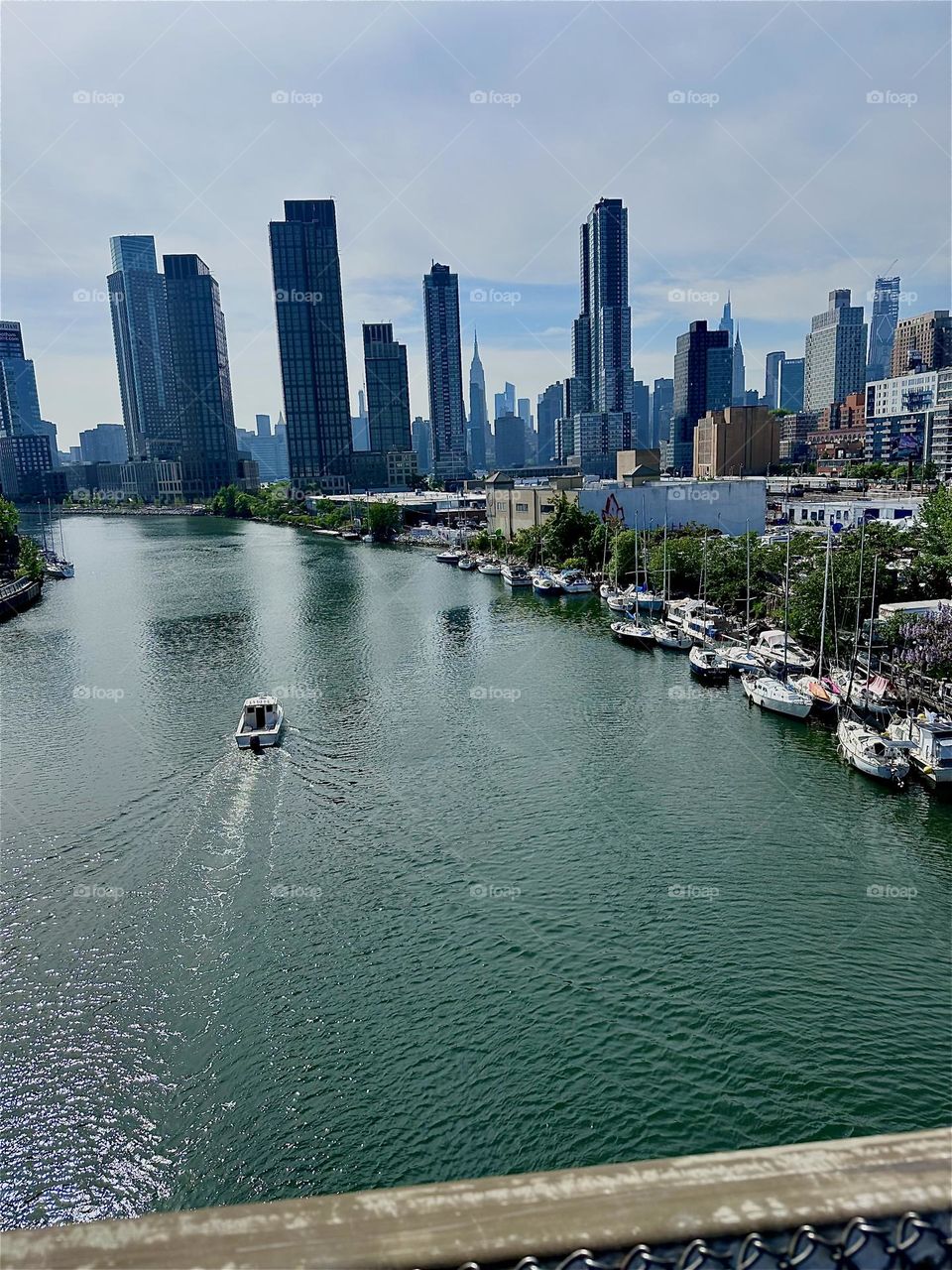This is picturesque “Newtown Creek” with its many boats seen from the “Pulaski Bridge” that connects LIC to “Greenpoint”, Bklyn. Across the “East River” in the distance we see “Manhattan” including the “Empire State Bldg”. 2024. Hypnotic Productions