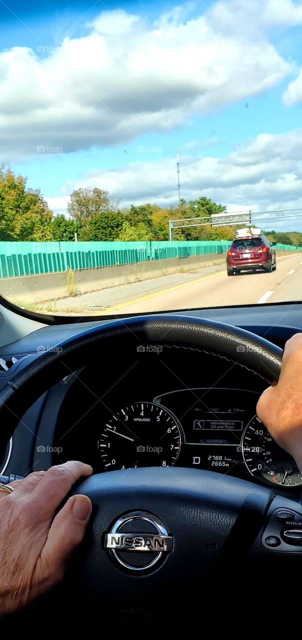 Hands on steering wheel showing dashboard and road ahead, blue sky with clouds ahead.