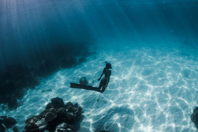 A female free diver underwater in crystal clear waters with light rays forming patterns on the sand. 