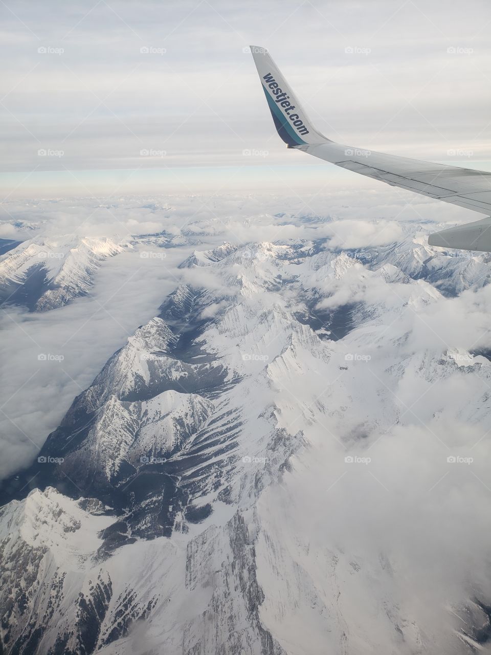 View from above, looking out of airplane window while flying over snow covered Canadian rockies during cloudy winter day.