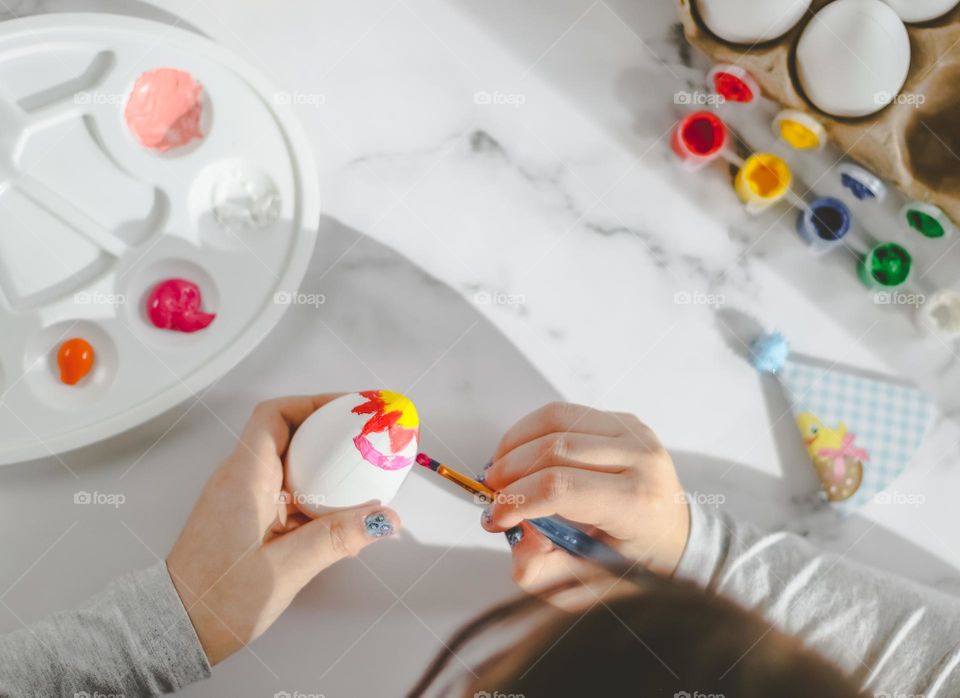 A little caucasian chewing gum draws with paints and a brush on a white egg, preparing for the celebration of Easter, sitting at a marble table, flat lay close-up. Concept preparation for Easter, children, diy.