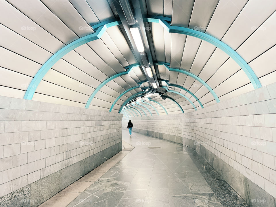 Tiny human in blue jeans walking by the grey-blue tunnel 