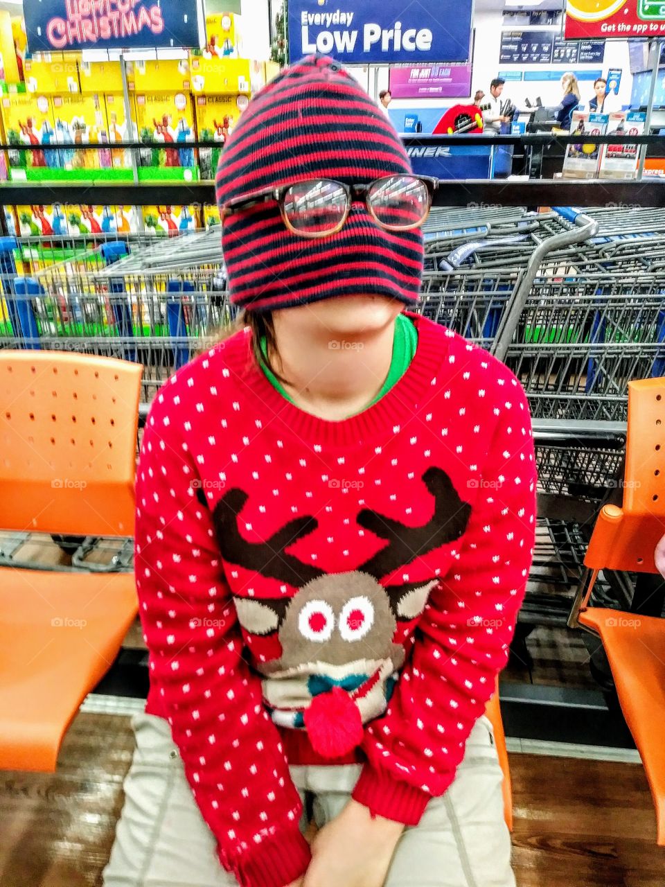 little girl in reindeer christmas sweater with beanie over face and glasses on beanie waiting in store chair