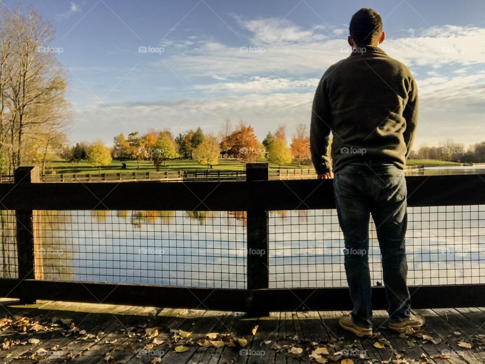 Male model standing on a footbridge by a lake on a beautiful autumn day with colorful trees in the background 