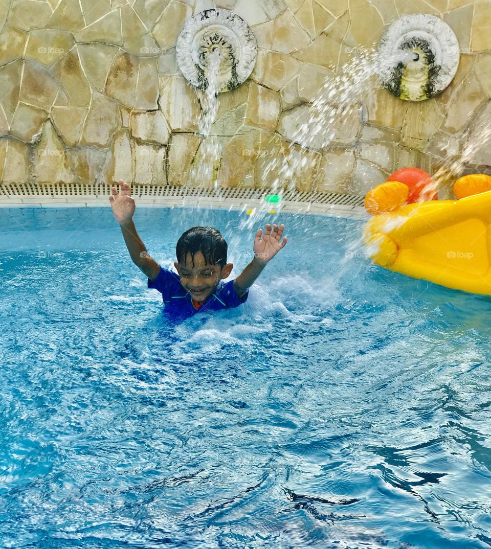 A 6 year old boy enjoying a lot at swimming pool in summer time and the boy is under water fountain and enjoying like anything he has a yellow floating duck.