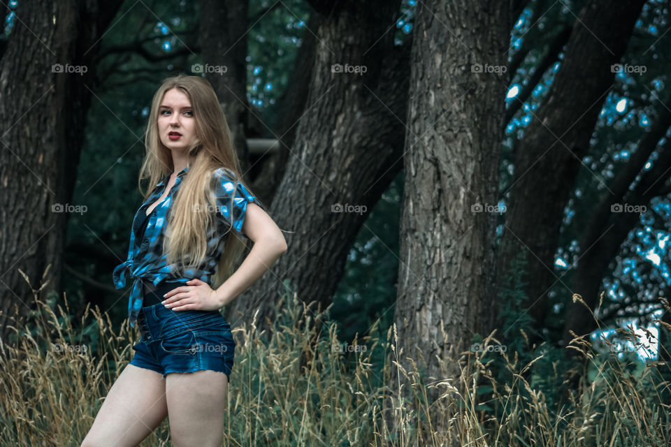 A girl with blond hair in a plaid shirt and short denim shorts on a background of trees and nature Girl, woman, man, people, blonde, blonde hair, checkered shirt, shorts shorts, denim shorts, forest nature, trees, grass, feelings, emotions tenderness, love, lifestyle, lifestyle, recreation