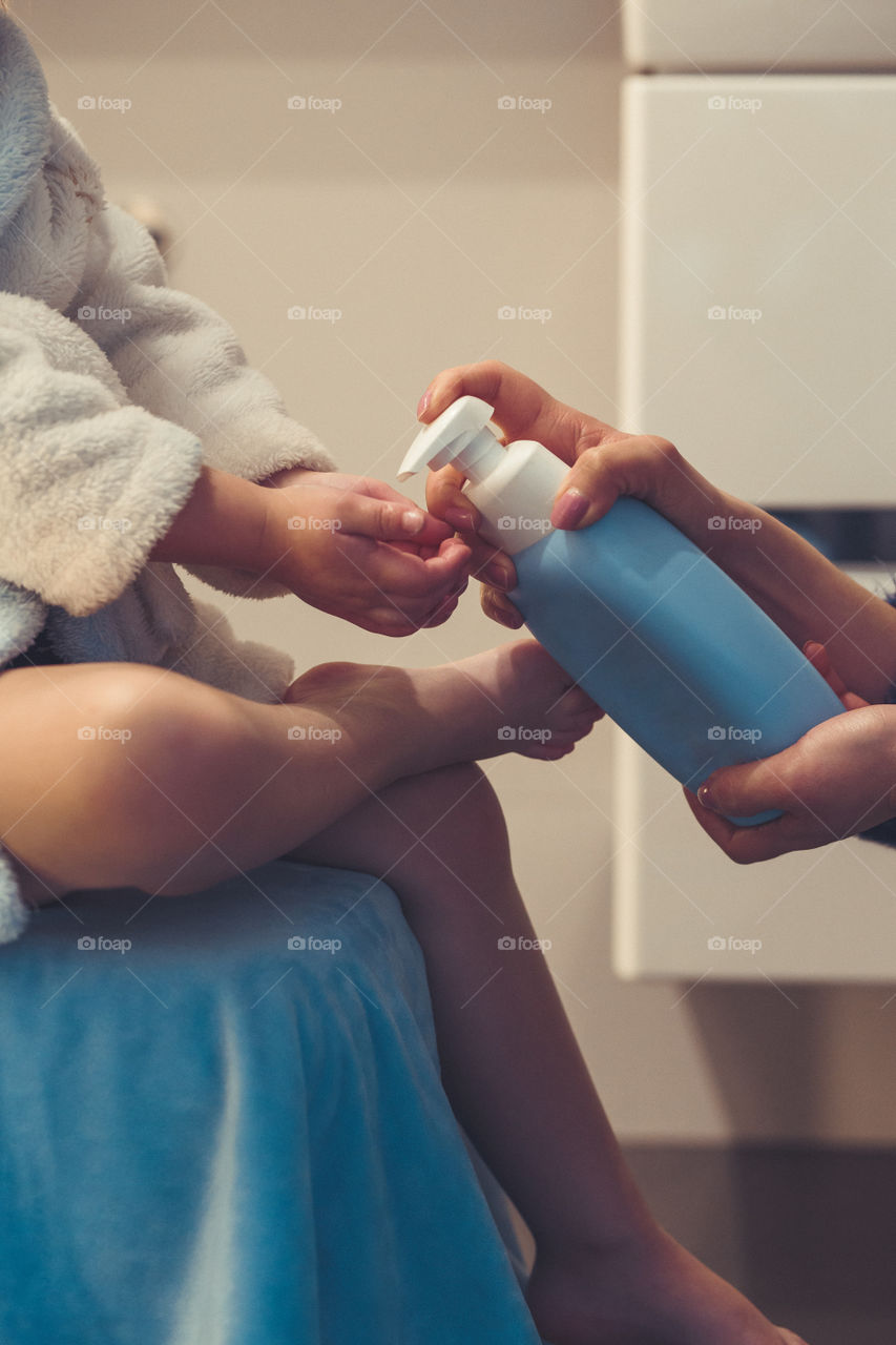 Mother applying moisturizing cream on her daughter's legs after bath. Mom caring about her child. Girl sitting in bathroom, wearing bathrobe. Real people, authentic situations