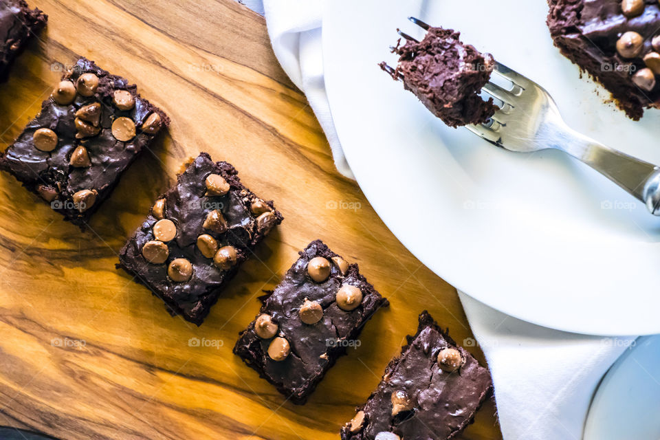 Rectangular, chocolate chip brownies placed on a wooden board with a white tablecloth and a fork on the side.