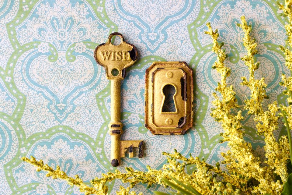 Flat lay of a rustic, yellow, vintage key embossed with the word “WISH,” keyhole, and dried yellow flowers on a light blue and green patterned background in natural light 