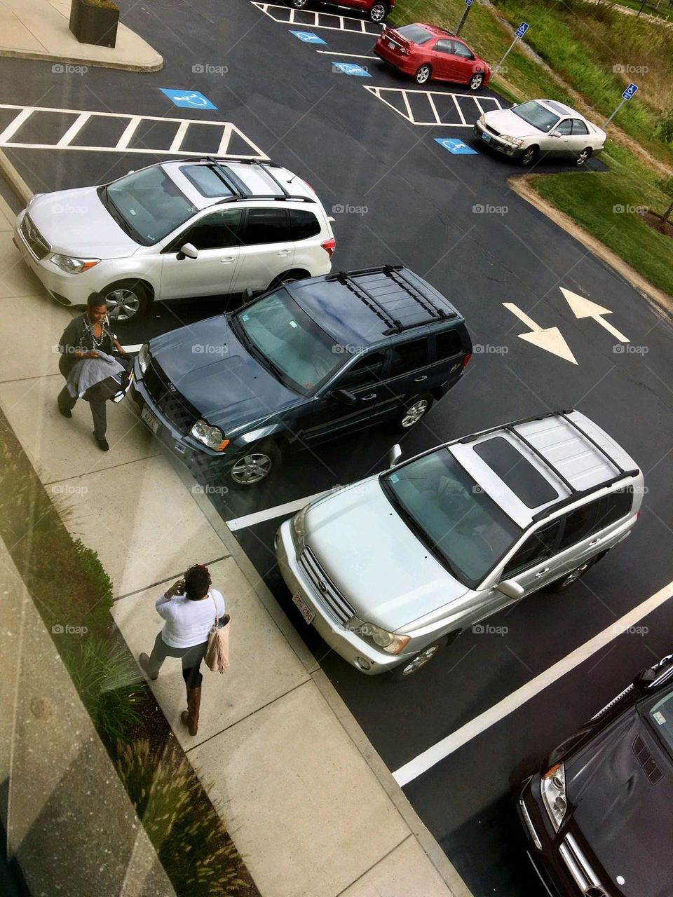 Parking lot full of cars near office building. People are walking to and from large doorway. The parking lot is well marked and painted. View is from the 2nd floor of building.