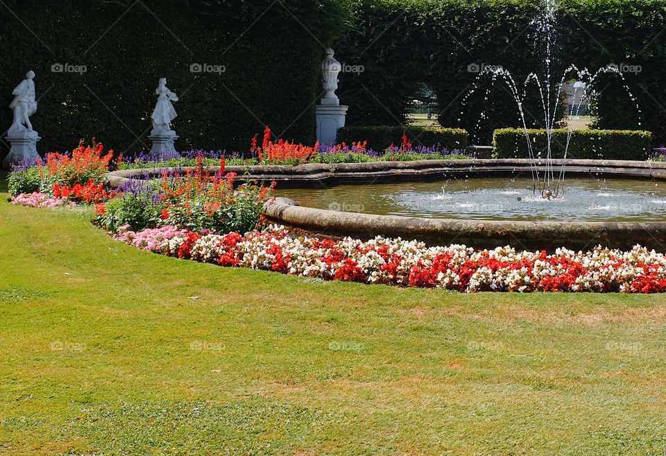 A beautiful outdoor fountain surrounded by bright red, pink, white, and purple flowerbeds along with statues and finely manicured hedges in the background in a public park in Europe on a sunny summer day. 