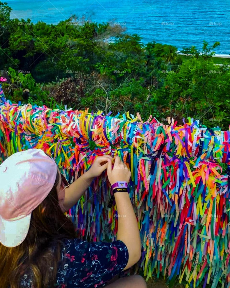 Teenager ties the ribbon of Senhor de Bomfim to the railing next to the Church. Superstition says that Bonfim's ribbons of different colors carry the power to fulfill three wishes. Porto Seguro Beach, Bahia Brazil, in the background.