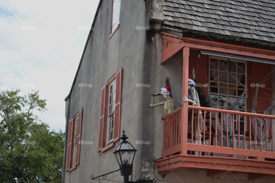 An old gray wooden house with red trimmed windows and balcony with pirate skeletons over looking the fence 