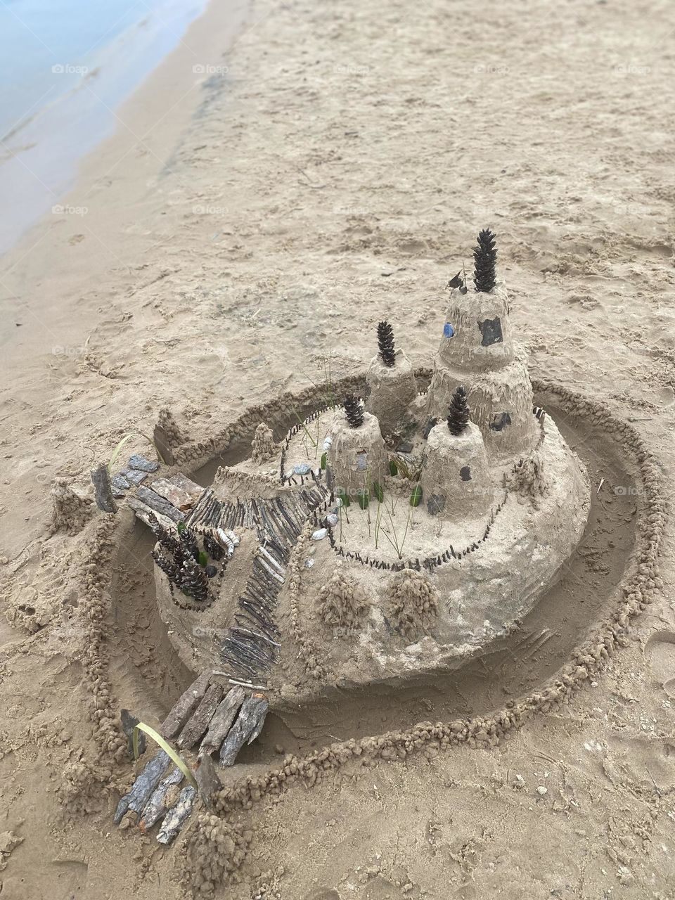 This image shows an intricate sandcastle on a beach, featuring multiple tiers and decorated with natural elements like pinecones, sticks, leaves, and shells. The sandcastle has a central tower surrounded by smaller structures, with detailed pathways 