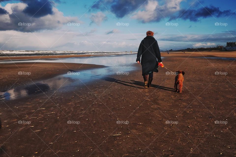 Woman walking on the beach with her dog on a beautiful sunny winter day, blue sky with clouds