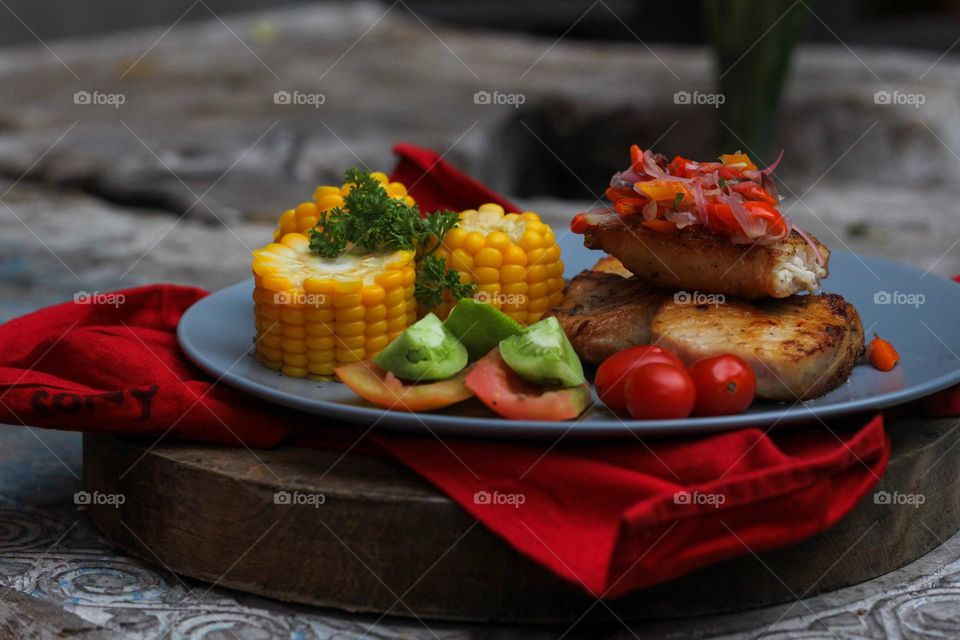 Tuna steak cooked with corn garnished with cherries, sliced tomatoes and lemon topped with sliced hot chili peppers on a baby blue melamine plate lined with red cloth and thick wood. Restaurant food.