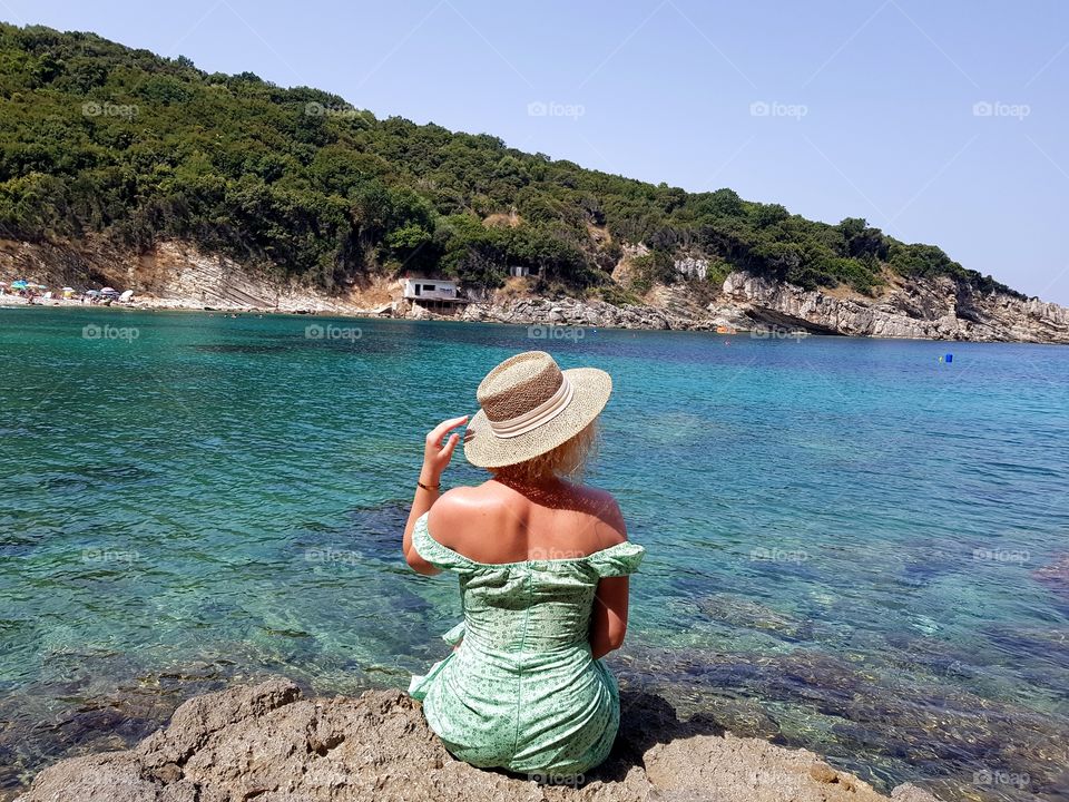 Close up back view of a woman sitting in a grren dress and hat looking out towards blue beach,ocean and sky. Isolated sea background for copy space. Summer holidays concept.