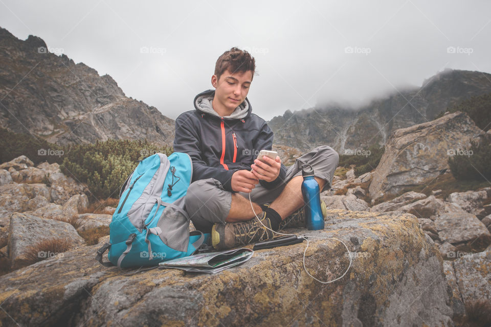 Boy resting on a rock and charging a mobile phone during the trekking in The Tatra Mountains