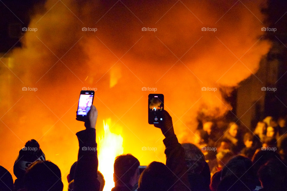 Crowd gathers around blazing tar barrel during demonstration, with on lookers filming with their cellphones.