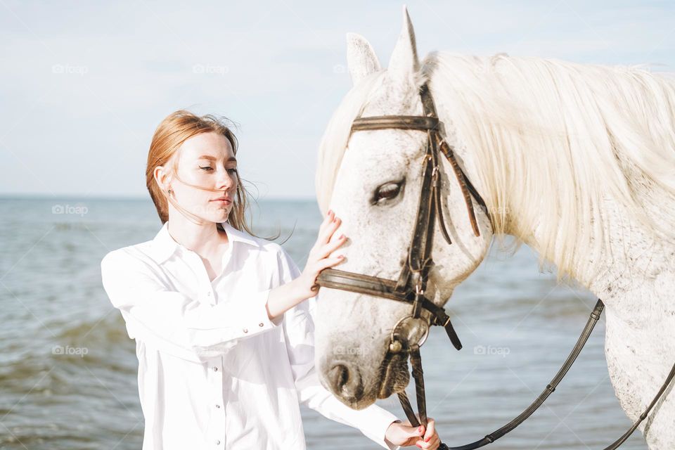 Young red haired woman in white shirt riding white horse on seascape background