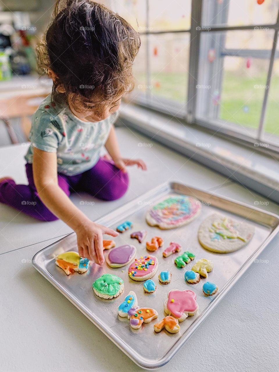 Toddler girl chooses sugar cookie to eat, toddler looks over sugar cookies she helped make with mother, proud toddler moments in the kitchen, Easter traditions in the kitchen, little girl helps make Easter sugar cookies