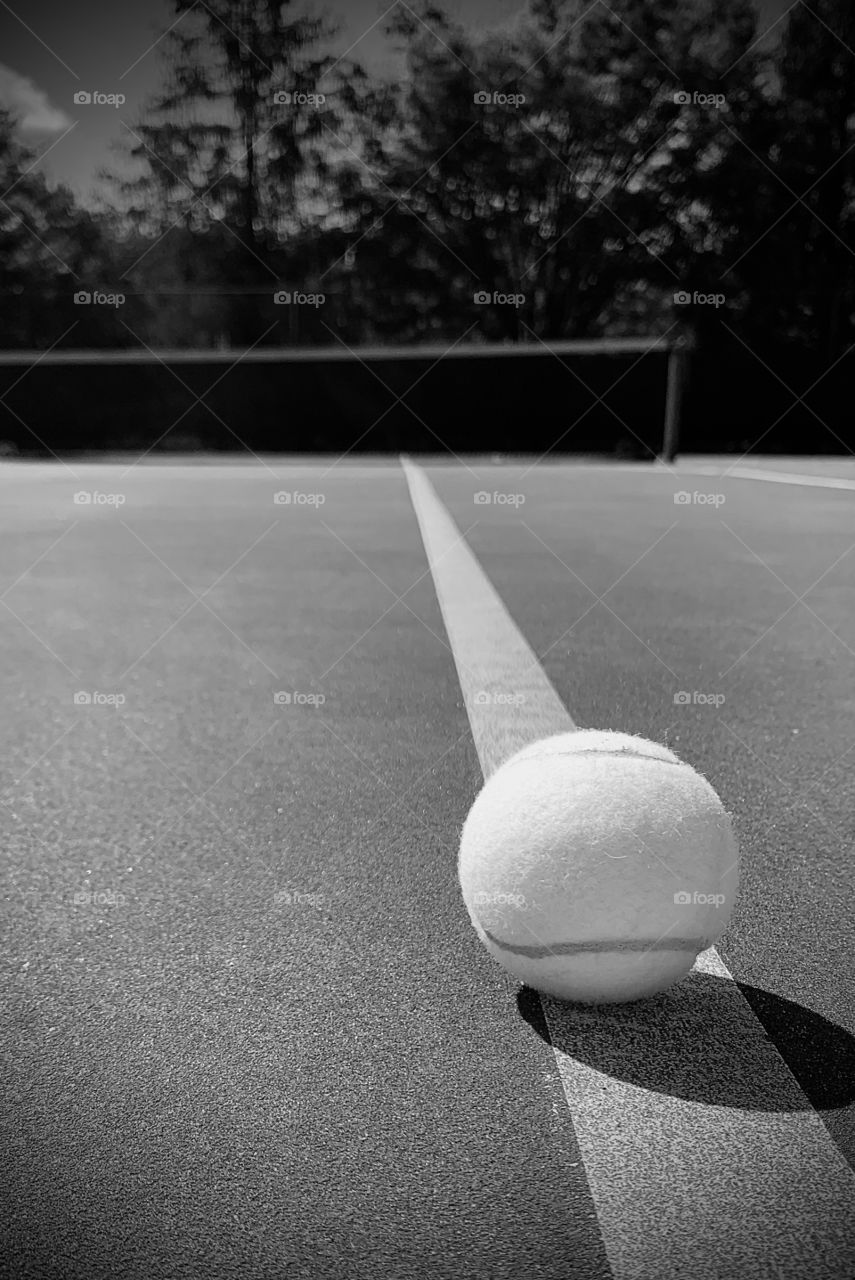 Black and white photo of a tennis ball on the line with a shadow. Looking out onto the court and net. 