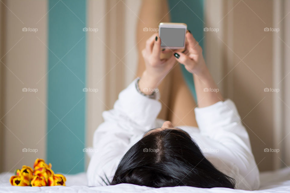 Woman in bed typing on phone with crossed legs up on the wall