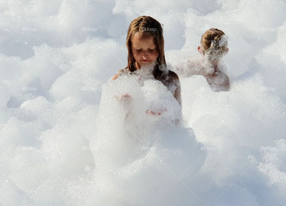 Two children covered in foam. One facing camera one standing with back at camera