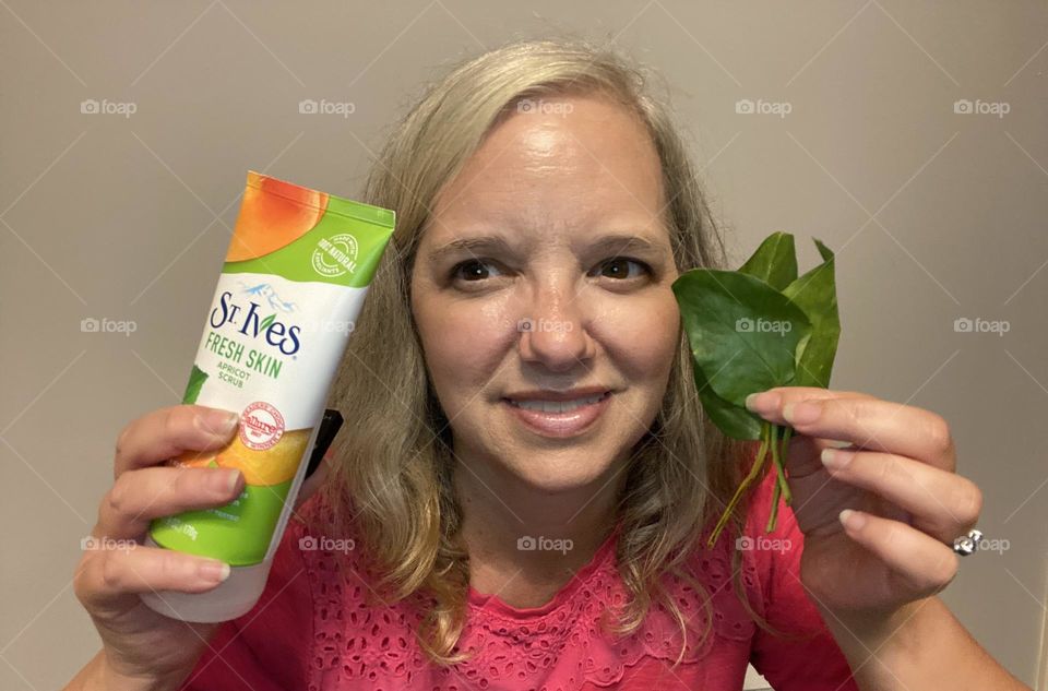 A woman (myself)holding St. Ives Fresh Skin Apricot Scrub in one hand and green plant leaves in the other. I love this product because it makes skin soft and has natural ingredients. Nature and this scrub go hand in hand.