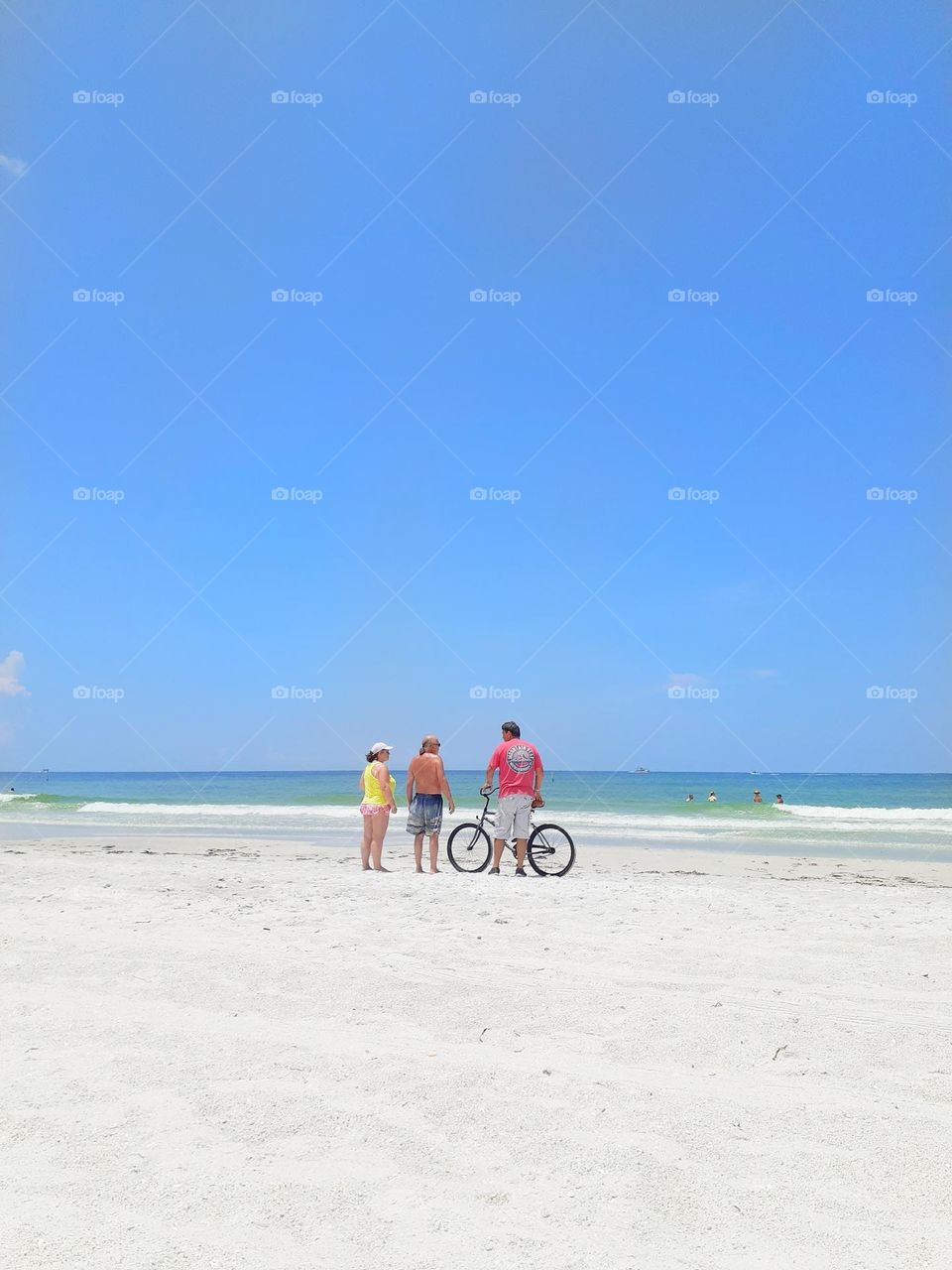 Three friends, two men and a woman stand on the beach talking. One of the men is holding a bicycle.