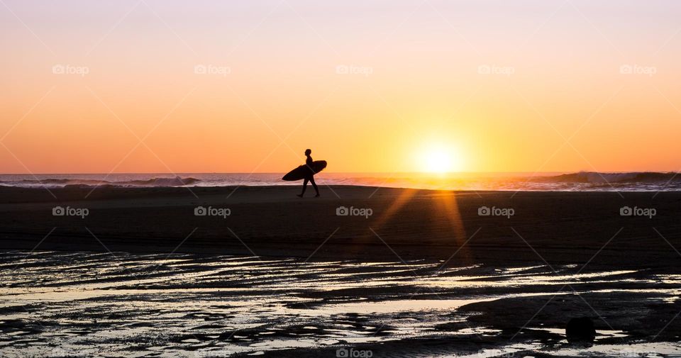 A surfer carrying a board is silhouetted by the sunset on the beach