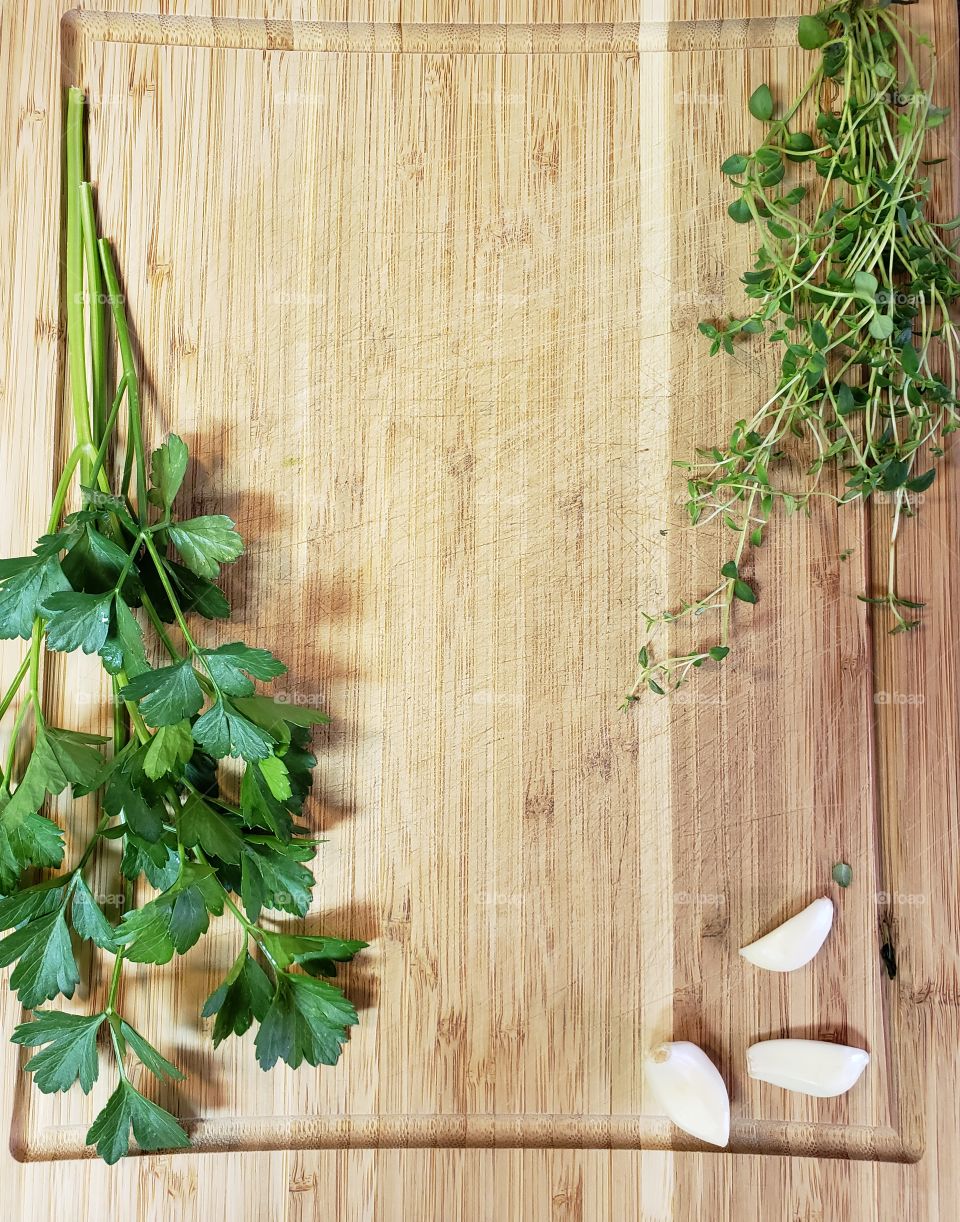 Fresh herbs of parsley, thyme, and garlic adorn a wooden light brown cutting board. 