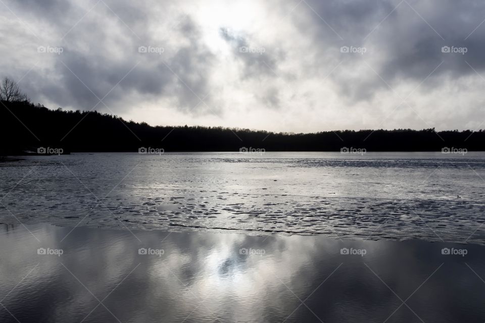 A dark grey day with dramatic clouds and reflection on the water. The ice on the lake is melting. Dramatiska moln och reflektion på sjön. Isen börjar smälta. Härlanda tjärn Sverige 