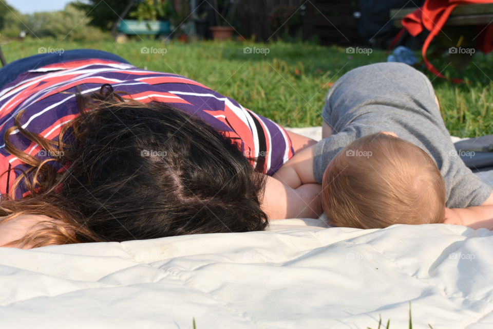 An older child and baby, siblings perhaps, lie on their tummies together, looking at each other lovingly, arms intertwined, bonding with each other. The older has dark hair with a red and purple shirt, the baby is in a gray onesie. 