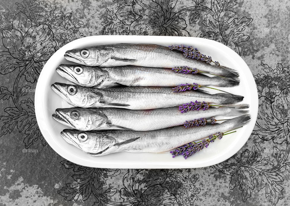 Fresh fish cleaned and prepared for cooking with sprigs of lavender on a white plate on a textured stone background. Freshly caught hake