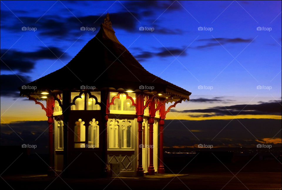 A Victorian seafront shelter is lit up at twilight in front of Bexhill beach, the lights of Eastbourne can be made out in the distance