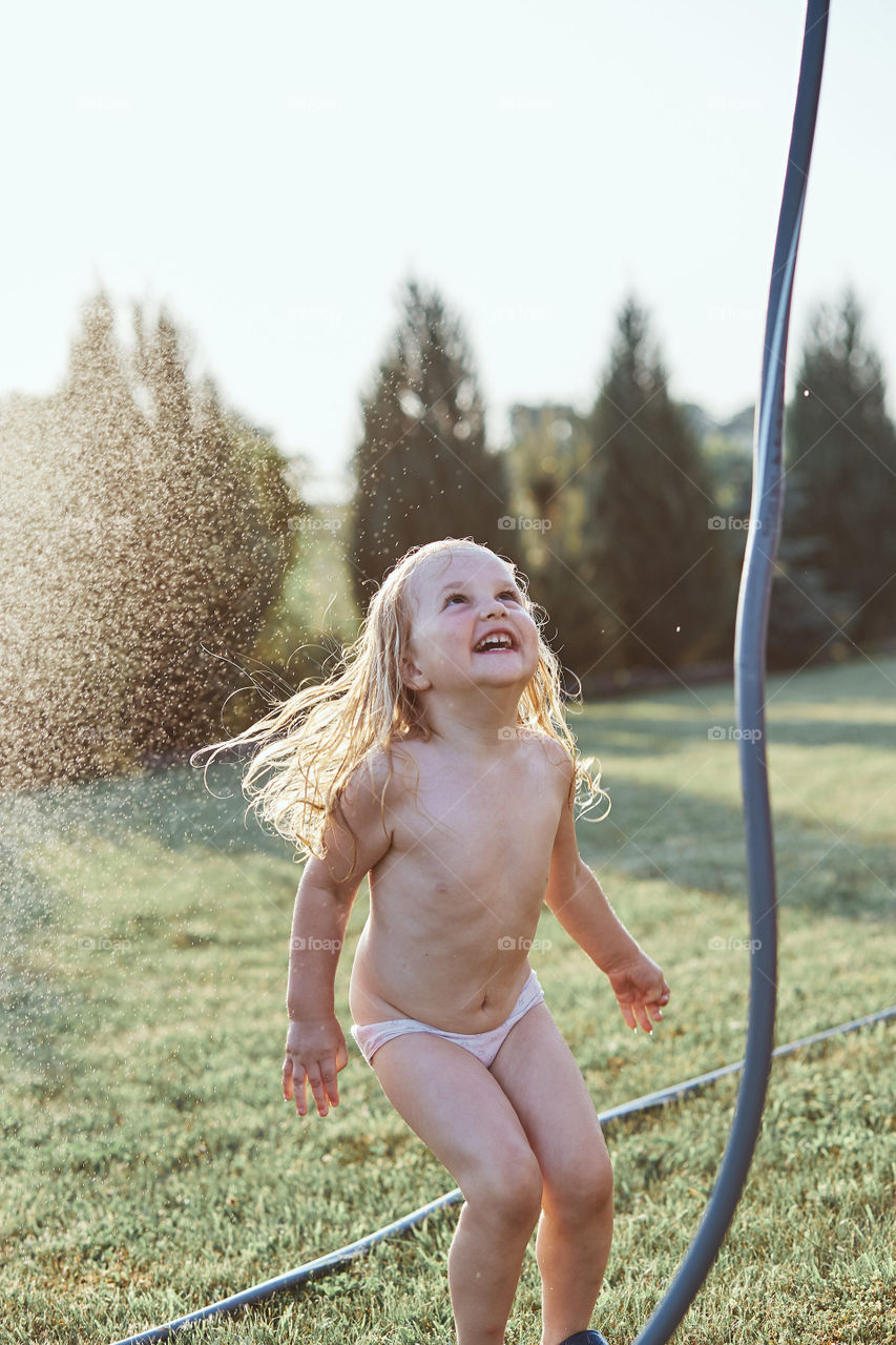 Little cute adorable girl enjoying a cool water sprayed by her mother during hot summer day in backyard. Candid people, real moments, authentic situations