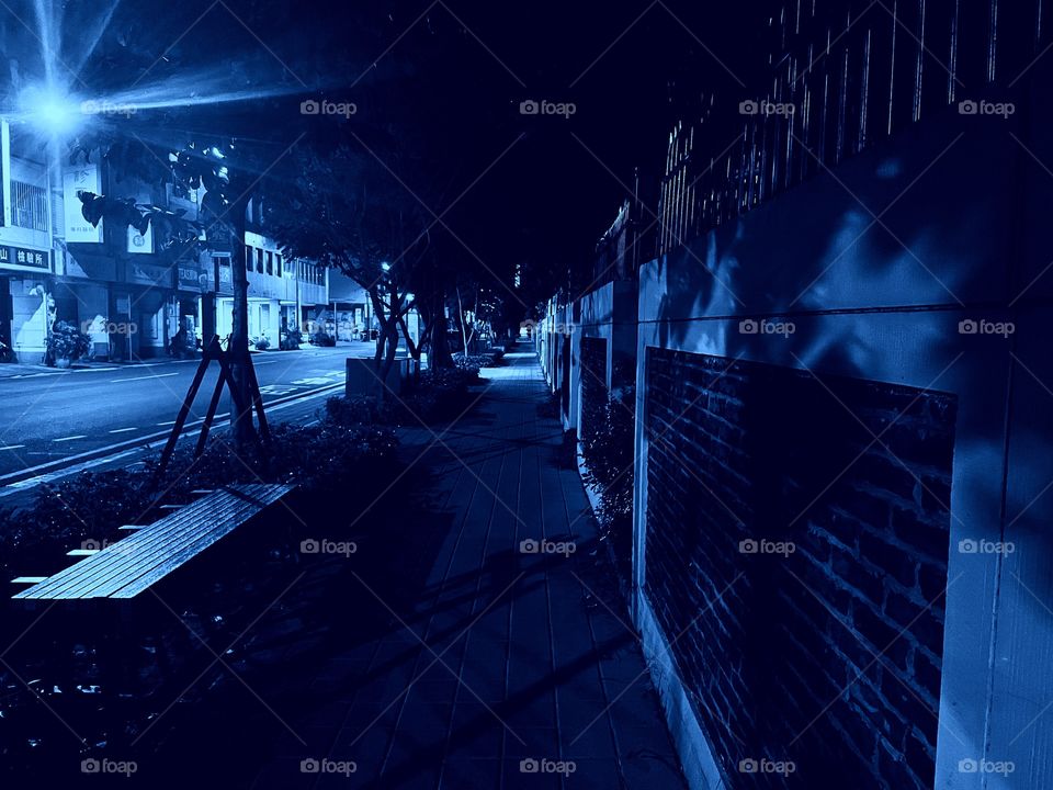 A nighttime photograph of a long, dark sidewalk. Several trees and benches are on the left, and a long brick wall to the right. 