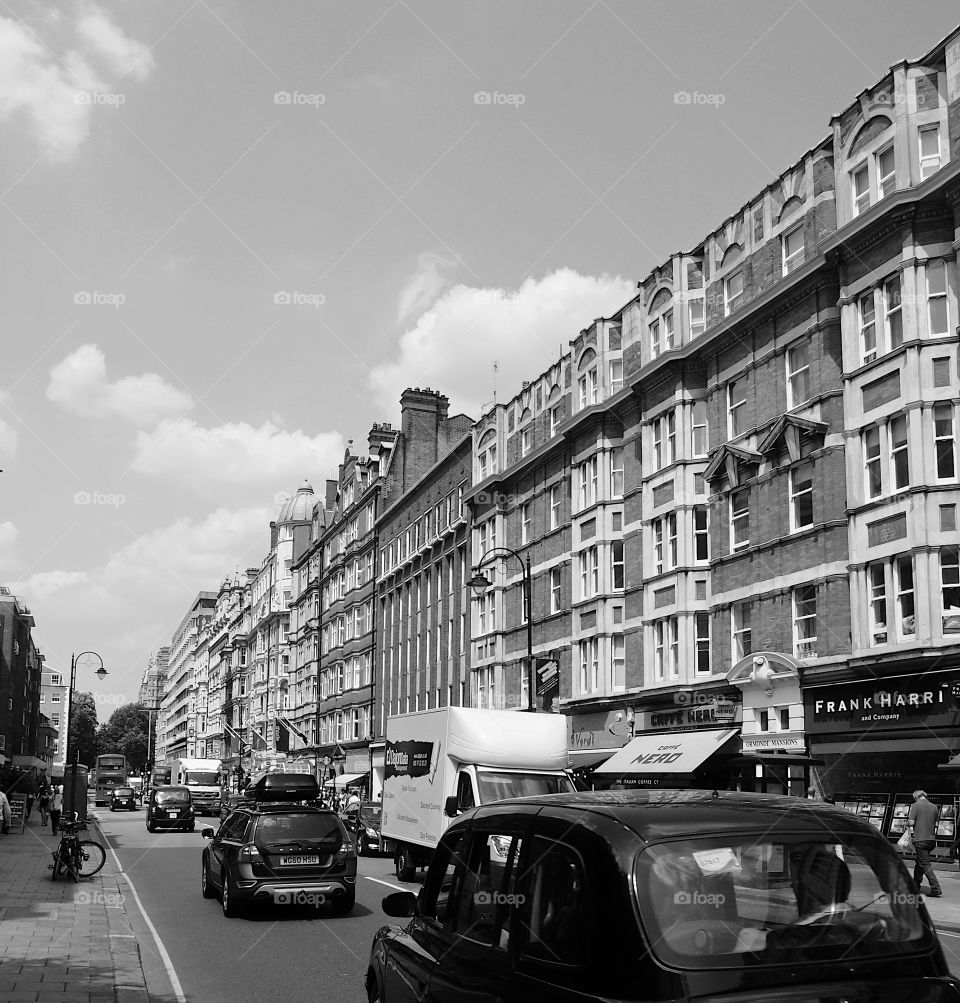 People walking and vehicles moving and parked on a busy ornate building lined buildings with shops on a busy street in London on a sunny summer day. 