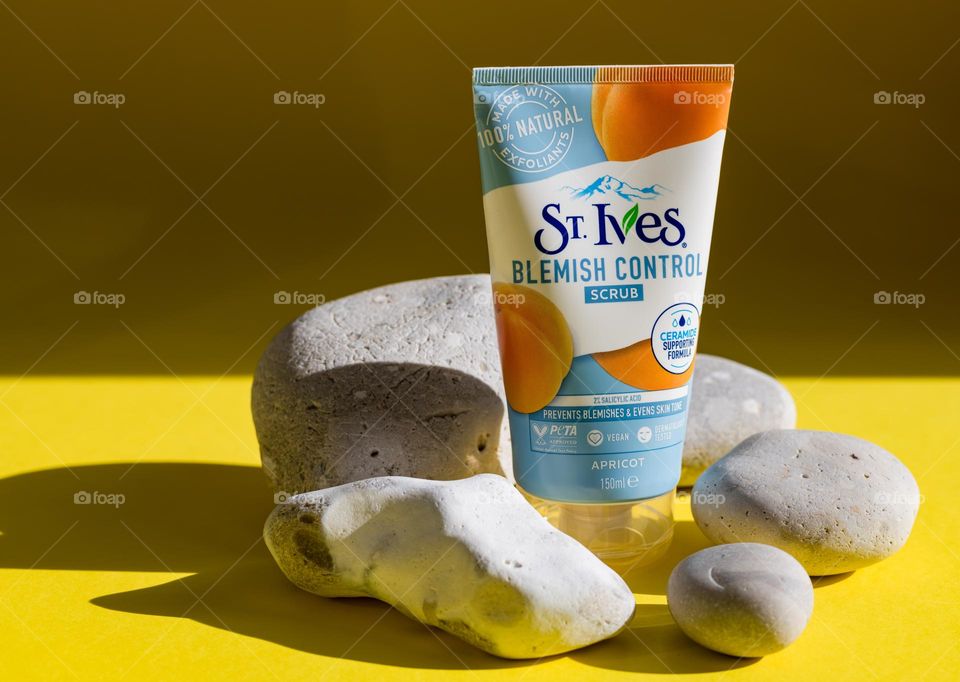 One tube of natural apricot scrub brand st. Ives stands on a yellow background surrounded by natural stones from the coast of the north sea with shadows and light from the sun, close-up side view. Concept of female cosmetics, body care, beauty.