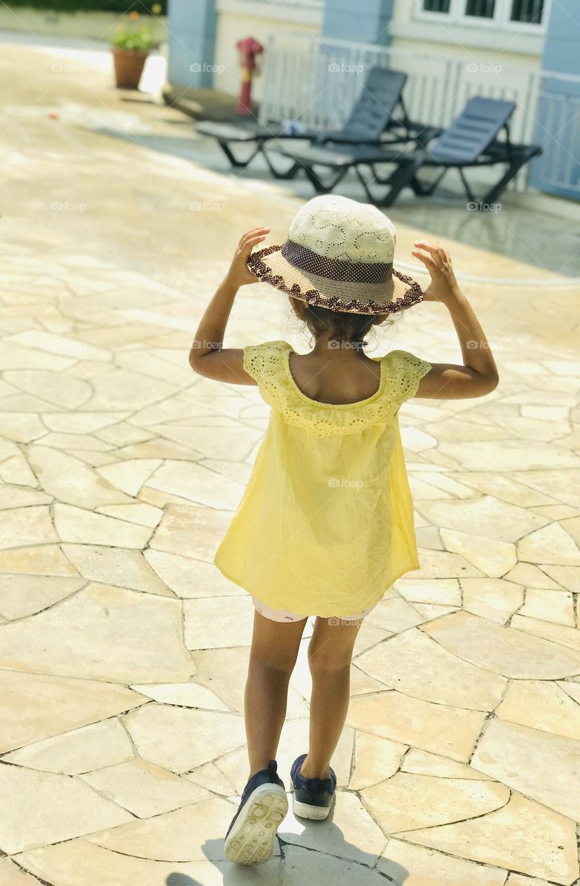 A baby girl with yellow summer outfit and hat on head going towards the swimming pool 