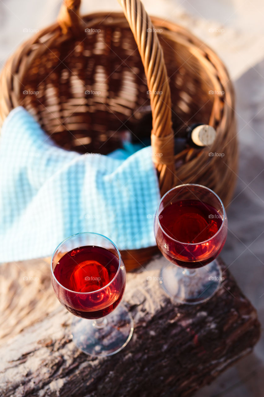 Two wine glasses with red wine standing on tree trunk, on beach, beside wicker basket with bottle of wine