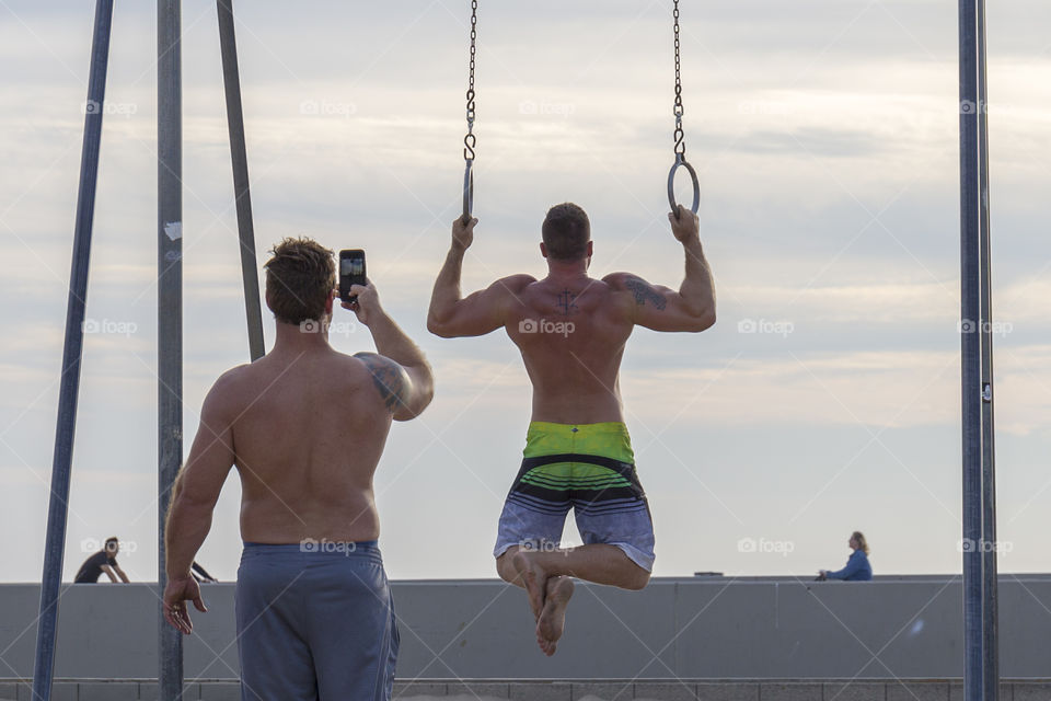 A man doing pull-ups with acrobatic rings, friend taking video 