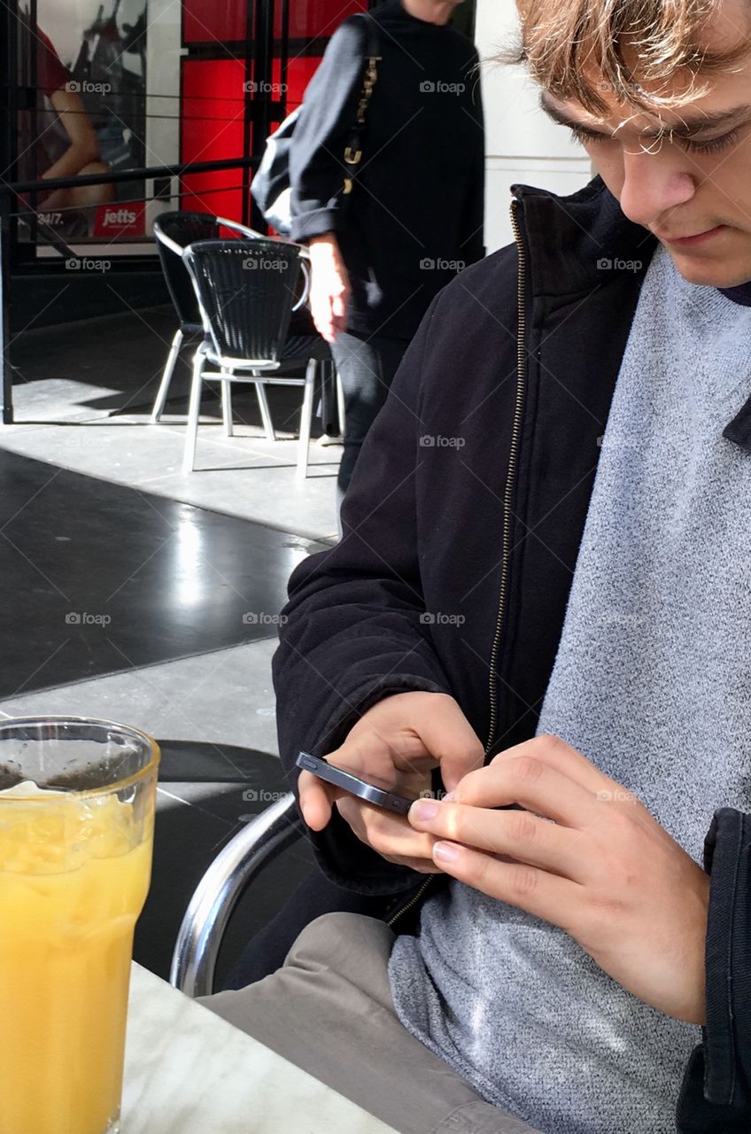 Young man sitting at outdoor cafe with glass of orange juice and using mobile phone